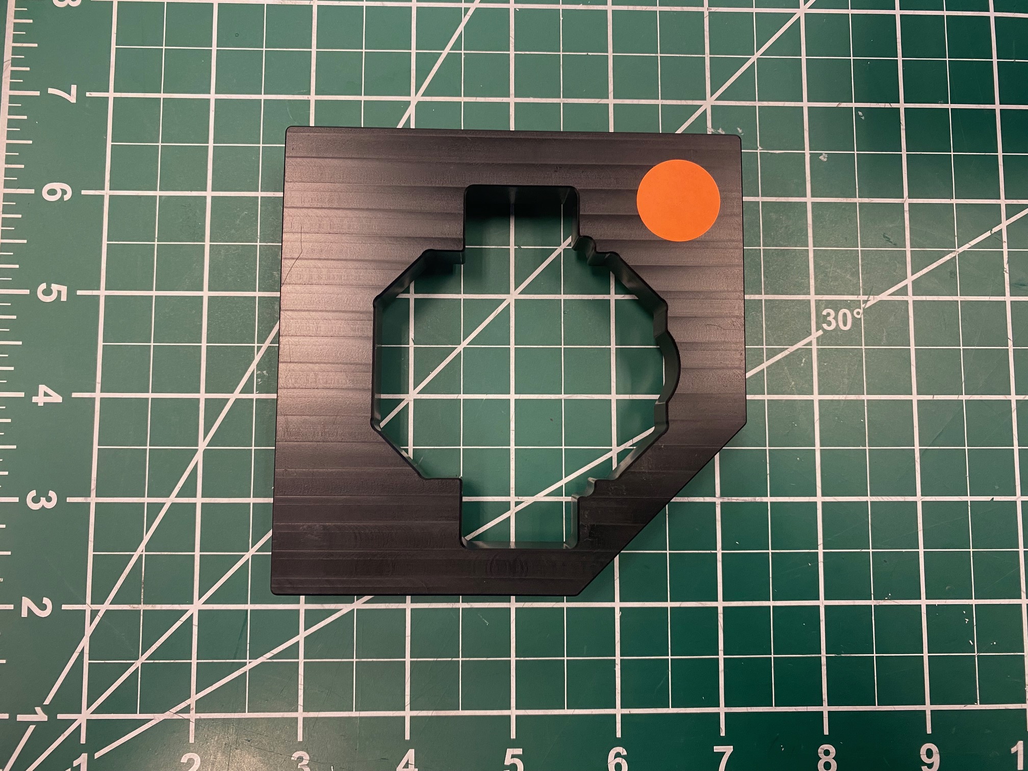 MAXSwerve Calibration tool with an orange dot indicating the side of the cutout that the bevel gear slots into. This part of the cutout is a more complex shape with more corners and curves than the other half of the cutout. 